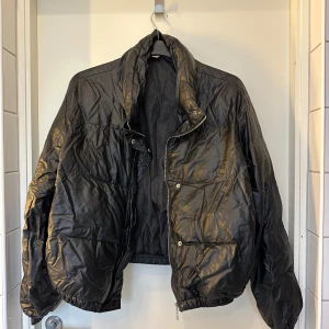 Svart pufferjacka i skinnlook från Flm - Cool svart pufferjacka från Flm med glansig skinnlook. Jackan har hög krage, dragkedja framtill och tryckknappar. Resår vid ärmslut och midja ger en schysst passform. Perfekt för dig som vill sticka ut med en edgy vibe. Hål i fickan som lätt kan sys ihop om man vill