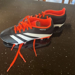 Adidas Predator fotbollsskor svart/vit - Säljer ett par Adidas Predator fotbollsskor i svart och vitt med orange detaljer och snörning. Skorna har klassiska tre ränder på sidan, låg profil och är designade för gräsplan. Insidan är fodrad i orange och yttermaterialet är syntet.