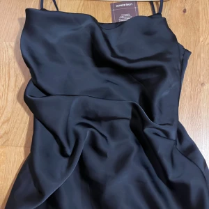 Mörkblå långklänning från Love Bonito - Säljer en elegant mörkblå långklänning från Love Bonito med tunna axelband och slät, glansig yta. Klänningen har en enkel och stilren design i ett mjukt, satinkänsligt material som faller snyggt längs kroppen. Perfekt för dig som gillar minimalistisk och chic stil.