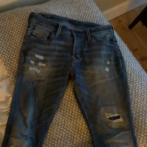 Blå slitna jeans med hål - Säljer ett par blå jeans med slitningar och hål på benen. De har klassisk femficksdesign och en mörk tvätt med ljusare partier. Passar dig som gillar en tuff och avslappnad stil. Perfekt till sneakers och hoodie.