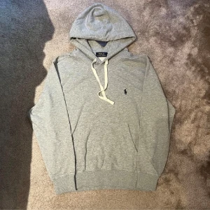 Grå hoodie från Polo Ralph Lauren - Säljer en stilren grå hoodie från Polo Ralph Lauren med klassisk huva och vita snören. Tröjan har en broderad logga på bröstet och en stor ficka framtill. Perfekt för chill dagar och enkel att matcha med jeans eller joggers.