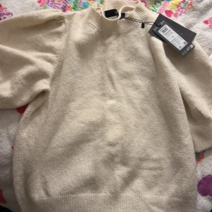 Beige stickad tröja Vero Moda XS - Superfin beige stickad tröja från Vero Moda med puffiga halvlånga ärmar och ribbad halsringning. Tröjan har en mjuk och mysig känsla, perfekt för att addera lite stil till din garderob. Snygg att matcha med jeans eller kjol.