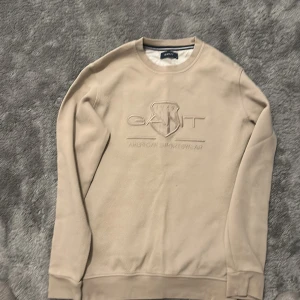 Beige sweatshirt från GANT i bomull - Snygg beige sweatshirt från GANT med broderad logga och sköld på bröstet. Tröjan har rund hals, ribbade muddar och är tillverkad i mjuk bomull. Perfekt för dig som gillar stilren och klassisk design med en sportig touch.
