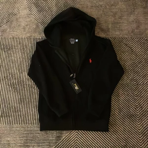 Svart hoodie från Polo Ralph Lauren - Svart hoodie från Polo Ralph Lauren i slim fit med dragkedja framtill och klassisk huva. Tröjan har det ikoniska röda Polo-logot på bröstet och är tillverkad i mjukt material. Perfekt för en chill och stilren look. Helt ny säljer den då den inte sitter på och har aldrig använt den som ni säkert ser på bilden. Skriv dm vid frågor och funderingar!