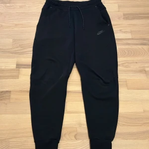 Nike tech svart - Svarta mjukisbyxor från Nike med dragsko i midjan och ribbade muddar vid bensluten. Byxorna har en diskret Nike-logga på framsidan och en praktisk ficka med dragkedja på baksidan. Perfekta för chill eller träning.