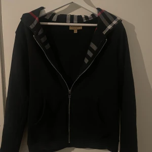  Burberry zip  hoodie - Svart hoodie från Burberry med klassiskt rutig foder i huvan i svart, vitt och rött. Tröjan har dragkedja framtill, broderad Burberry-logga på bröstet och känguruficka. Perfekt för dig som vill ha en stilren och lyxig streetwear-look. Pris kan diskuteras 