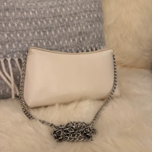 Beige axelväska med kedja - Säljer min beiga väska som jag hade på min bal. Använd en gång alltså. Bytte kedjan själv mot en silvrig så finns två gulddetaljer kvar men som inte syns. Dels kan man använda kedjan som en axelväska men man kan även stoppa in kedjan och få det till en clutchbag. Passar till alla färger på klänningar, skor och smycken!