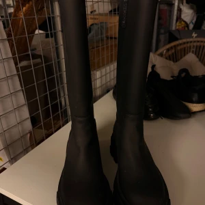 Svarta höga boots med platt sula - Säljer ett par svarta höga boots med platt sula och minimalistisk design. Skorna har en rund tå och är tillverkade i ett slätt material som ger en clean look. 