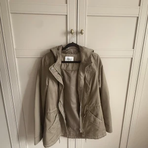 Grön parkajacka från ONLY med huva - Snygg grön parkajacka från ONLY med praktisk huva, dragkedja och knappar framtill. Jackan har två stora fickor med lock och snörning i midjan för att justera passformen. 