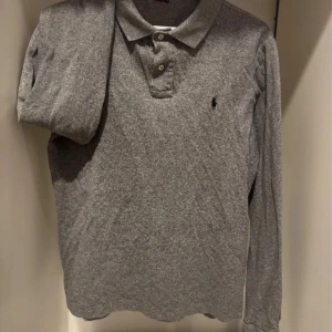 Grå långärmad pikétröja Polo Ralph Lauren - Snygg grå långärmad pikétröja från Polo Ralph Lauren i slim fit-modell. Klassisk krage med knappar och ikoniska broderade loggan på bröstet. Perfekt för dig som gillar stilren och enkel design. Tillverkad i mjuk bomull för skön känsla.