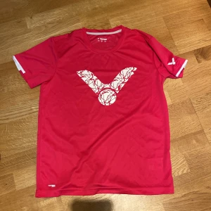 Victor - Röd sportig t-shirt - Badminton  - Snygg röd t-shirt från Victor med stort grafiskt vitt tryck på bröstet. T-shirten har rund hals, korta ärmar och är gjord i ett lätt polyester-material som andas. Perfekt för dig som gillar att träna och vill ha en cool look på gymmet eller på planen.