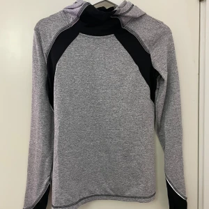 Grå och svart tränings tröja med tumhål - Sportig tröja i grått och svart med långärmad design och tumhål vid ärmsluten. Tröjan har huva och kontrastpaneler i svart på bröst, ärmar och huva. Tillverkad i mjukt syntetmaterial som andas, perfekt för träning eller chill. Snygga sömmar ger en modern vibe.