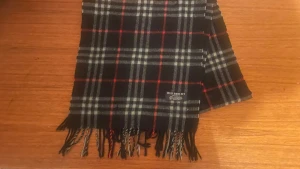 Rutig svart halsduk från Burberry - Klassisk halsduk från Burberry i 100% kashmir. Svart botten med rutigt mönster i vitt, grått och röda linjer. Tidlös design med fransar i ändarna och diskret Burberry-märke. Perfekt accessoar för att lyfta din outfit.