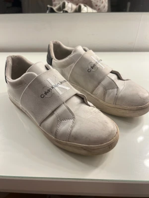Vita Calvin Klein Jeans sneakers - Snygga vita sneakers från Calvin Klein Jeans med bred elastisk rem över foten och tydligt CK-logga. Skorna har rund tå, platt sula och svart detalj på hälen med logotyp. Det är även bandet smash into pieces autograf på ena skon