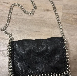 Svart axelväska med kedja från Tiamo - Snygg svart axelväska från Tiamo i skinn med grov silverfärgad kedja som axelrem. Väskan har ett coolt flätat kantband och magnetlås. Insidan har dragkedjefack för småsaker. Perfekt storlek för det viktigaste och ger en edgy vibe till din outfit.