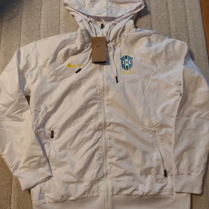 Vit Brasil Nike vindjacka med huva - Snygg vit vindjacka från Nike med Brasil CBF-logga på bröstet och gul swoosh. Jackan har huva med snörning, hel dragkedja och två sidofickor med dragkedja. 📏Storlek:Medium 📦Skick:Helt ny 👕Endast jackan ingår"