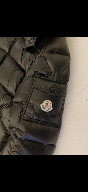 Svart Moncler dunjacka med huva - Säljer en svart dunjacka från Moncler med glansig finish, huva och dragkedja framtill. Jackan har Moncler-logga på ärmen, stora fickor med dragkedja och en cool serietidningsetikett på insidan. Perfekt för kalla dagar när du vill hålla stilen.