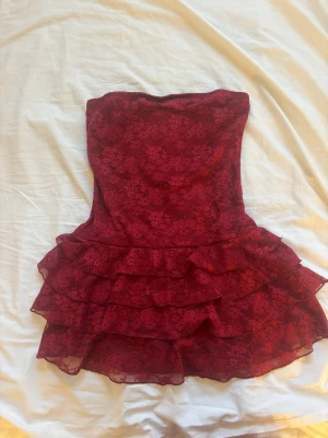 Vinröd axelbandslös spetsklänning - Säljer en vinröd axelbandslös klänning med flera lager volanger och vacker spets över hela klänningen. Den har en kort och figurnära passform som ger en snygg siluett. Perfekt för dig som vill sticka ut med en romantisk och trendig look.