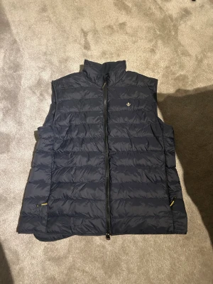 Mörkblå dunväst från Morris - Snygg mörkblå dunväst från Morris med quiltad design och hög krage. Västen har dragkedja framtill, två fickor med dragkedja och en liten broderad logga på bröstet. Perfekt för lager-på-lager och stilren look.