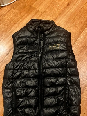 Svart dunväst från EA7 Emporio Armani - Snygg svart dunväst från EA7 Emporio Armani med guldfärgad logga på bröstet. Västen har quiltad design, hög krage och dragkedja framtill med EA7-detalj. Perfekt för lager-på-lager och streetstyle. Inga defekter eller skador. Kom pm för mer info 