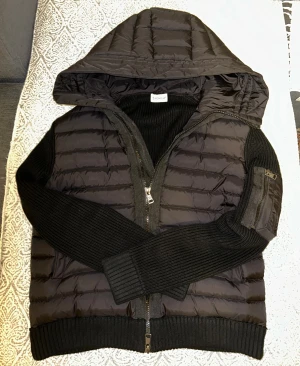 Svart Moncler pufferjacka med huva - Svart pufferjacka från Moncler med stickade ärmar och ribbade muddar. Jackan har huva, tvåvägsdragkedja och en ficka med dragkedja på ärmen. Materialmix av polyester och stickad ull ger en schysst look. Perfekt för dig som gillar streetstyle.