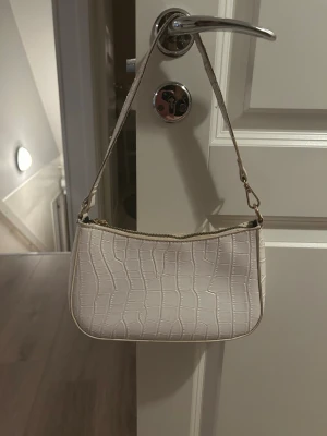 Vit axelväska med krokodilmönster - Snygg vit axelväska med krokodilmönstrad yta och guldfärgade detaljer. Väskan har en stilren form och dragkedja upptill. Perfekt för att lyfta din outfit med en trendig touch. Materialet är syntet och remmen är justerbar.