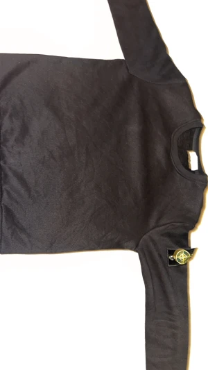 Svart långärmad Stone Island tröja - Svart långärmad tröja från Stone Island med klassisk rund hals och ikonisk patch på vänster ärm. Tröjan är i mjuk bomull och har en clean, stilren look som passar perfekt till jeans eller cargo. Snyggt ribbad vid halsen för extra komfort.