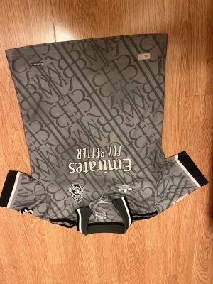 Grå Real Madrid fotbollströja Adidas S - Säljer en grå Real Madrid fotbollströja från Adidas med tryckta loggor och mönster över hela tröjan. Nummer 24 och namnet 'Arda Güler' på ryggen. Kortärmad med svart och vit ribbad krage och ärmslut. Materialet är lätt och andas, perfekt för match eller träning.