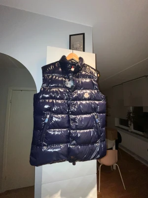 Mörkblå dunväst från Moncler - Snygg mörkblå dunväst från Moncler med glansig finish med puff-design. Västen har hög krage, två dragkedjefickor och ikonisk Moncler-logga på bröstet. Materialet är nylon och dun, perfekt för kalla dagar. Stängs med dragkedja och tryckknappar framtill. Inte använd så all nödvändig plast är kvar! Pris kan diskuteras vid snabb affär!