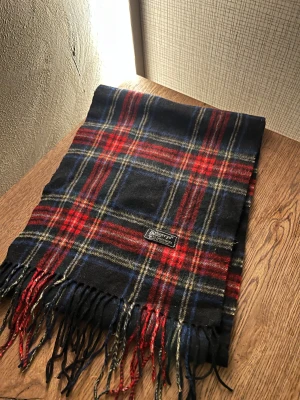 Rutig ullhalsduk från Burberrys - Klassisk rutig halsduk från Burberrys i mjuk ull. Färgerna är svart, röd, blå och inslag av beige och vitt. Halsduken har fransar i ändarna och en tidlös design som ger en cool och stilren vibe.