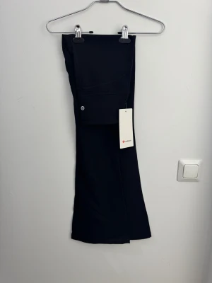Svarta bootcut/flared leggings/tights från Lululemon storlek 4 - Svarta leggings från Lululemon med hög midja och diskret logga på benet. Tillverkade i supermjuk och stretchig Nulu™-mix med Lycra för extra komfort. Byxorna har dold ficka i linningen och är perfekta för yoga eller chill. Snygg och enkel design.