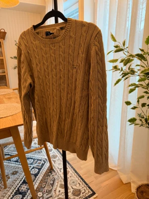 Beige kabelstickad tröja från GANT - Snygg beige kabelstickad tröja från GANT i premium bomull. Klassisk rund hals och ribbade muddar vid ärmslut och nederkant. Perfekt för dig som gillar en clean och tidlös look. Liten GANT-logga broderad på bröstet. Passar dig som vill ha en normal passform.