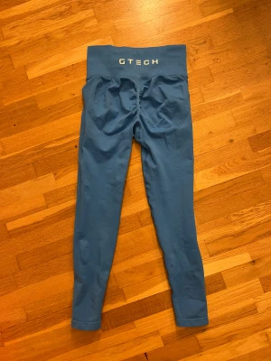 Blå höga leggings från GTECH - Snygga blå leggings från GTECH med hög midja och ribbad linning. Materialet är stretchigt och har mesh-detaljer längs benen för extra ventilation. Logga på linningen och framtill. Perfekta för gymmet eller annan träning.