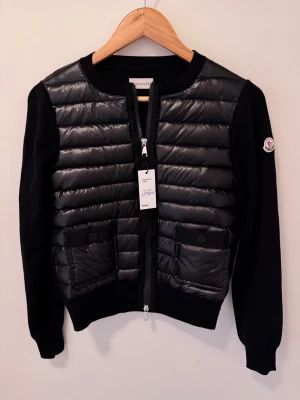 Moncler | women cardigan - Svart cardigan jacka från Moncler med quiltad front i blankt material och stickade ärmar. Jackan har två fickor med knappstängning, rund hals och dragkedja framtill med Moncler-logga. Perfekt för dig som gillar stilren och sportig vibe.