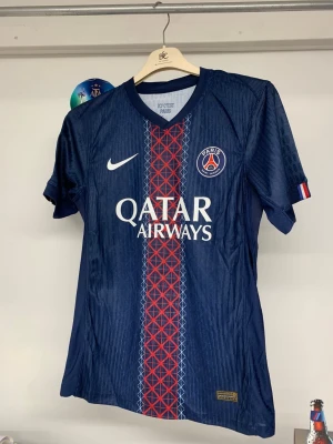 PSG Dembele #10 Champions League tröja - Paris Saint-Germain matchtröja med O. Dembélé och nummer 10 på ryggen. Mörkblå med rött och vitt geometriskt mönster längs mitten, PSG-logga, Nike Swoosh och Qatar Airways tryck. Champions League och UEFA Foundation patch på ärmen. Tillverkad i lätt, ventilerande material.