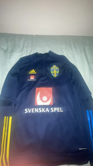Svenska landslaget långärmad Adidas tröja - Säljer en mörkblå långärmad fotbollströja från svenska landslaget, Adidas Aeroready. Tröjan har gula och blå ränder på ärmarna, landslagets emblem och Svenska Spel-logga på bröstet. Materialet är lätt och andas, perfekt för träning eller match.