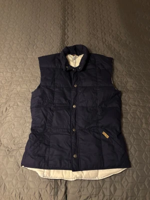 Mörkblå dunväst från Carina - Snygg mörkblå dunväst från Carina med hög krage och vita detaljer på insidan. Västen har tryckknappar framtill, två fickor och quiltad design. Perfekt för lager-på-lager och håller dig varm under kyliga dagar.