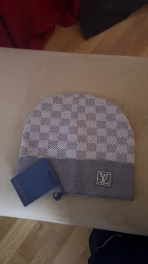 Grå rutig mössa från Louis Vuittons - Snygg grå mössa från Louis Vuitton med klassiskt rutigt mönster och LV-logga framtill. Mössan har en bred uppvikt kant och är stickad i mjukt material som håller dig varm och stilren. Perfekt accessoar för att lyfta din look.