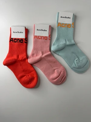 Färgglada Acne Studios strumpor - Tre par snygga strumpor från Acne Studios i rött, rosa och ljusblått. Strumporna har ribbade skaft och Acne Studios-logga upptill. Tillverkade i mjuk bomullsblandning för skön passform. Perfekta för att liva upp din outfit.
