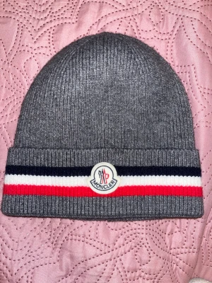 Grå  mössa med ränder - Snygg grå ribbstickad mössa med klassisk logga framtill. Mössan har bred uppvikt kant med marinblå, vit och röd rand som ger en sportig vibe. Perfekt för dig som vill ha både stil och värme under kyliga dagar.