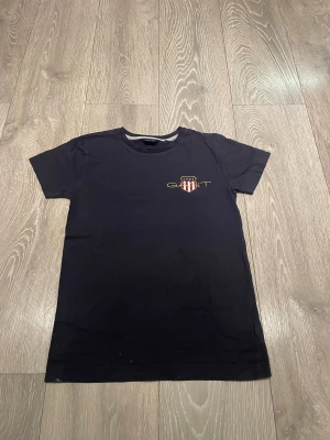 Mörkblå  GANT t-shirt med broderad logga - Mörkblå t-shirt från GANT med broderad logga och sköld på bröstet. Klassisk rund halsringning och korta ärmar. Tillverkad i mjuk bomull för en skön känsla. Perfekt basic med snygg detalj framtill. Osäker på storlek, troligen 130-140. Köpare betalar frakt. 
