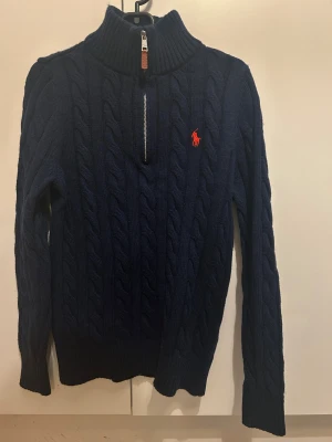 Marinblå stickad polotröja Polo Ralph Lauren - Snygg marinblå stickad polotröja från Polo Ralph Lauren med klassisk kabelstickning och röd logga på bröstet. Tröjan har half zip i halsen och ribbade muddar. Perfekt för dig som gillar stilrena och tidlösa plagg.