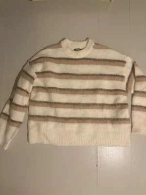 Randig fluffig tröja från Gina Tricot - Supermjuk och fluffig stickad tröja från Gina Tricot i vitt och beige med breda horisontella ränder. Rund hals och långa ärmar, perfekt för att hålla dig varm och snygg under kyliga dagar. Ribbad kant vid hals, ärmslut och nederkant.