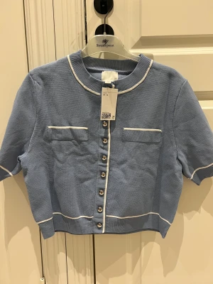 Blå stickad topp från HM - ljusblå stickad topp från hm med korta ärmar, vita kontrastkanter och silvriga knappar framtill. Två dekorativa fickor på bröstet och croppad passform. Aldrig använd 