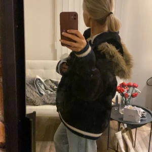 Svart teddyjacka med fuskpäls - SNYGG Svart vintagejacka från Sir Benni Miles med huva och fuskpäls i brunt runt kanten. Jackan har ribbade muddar med vita ränder, dragkedja framtill och två fickor. Perfekt för kalla dagar och har en loose passform. Jackan är väldigt varmt!!!😍