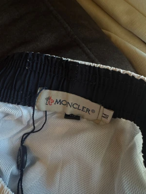 Vita Moncler shorts med logga - Snygga vita shorts från Moncler med broderad logga framtill. De har elastisk midja med snörning och är tillverkade i ett lätt syntetmaterial med meshfoder. Perfekta för sommardagar när du vill ha en clean och exklusiv look.