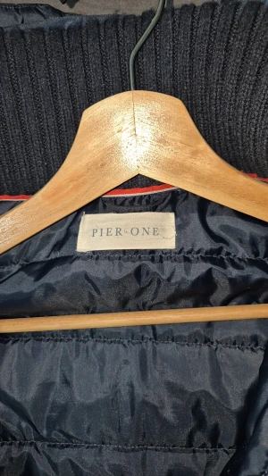 Mörkblå pufferjacka från Pier One - Snygg mörkblå pufferjacka från Pier One med stickade ärmar och krage. Jackan har två dragkedjefickor framtill, quiltad kropp och ribbade muddar. Perfekt för kyliga dagar och har en sportig vibe med diskret röd detalj vid kragen.