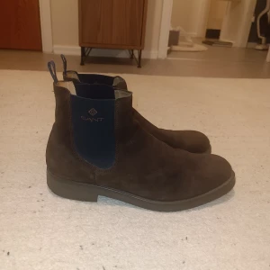 Bruna Chelsea boots från Gant - Snygga Chelsea boots från Gant i mörkbrun mocka med elastiska blå paneler på sidorna och dragflikar med logga. Rund tå och platt sula ger en klassisk och stilren look. Perfekta för dig som gillar tidlös design och vill ha boots som funkar året runt.