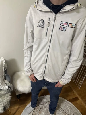Vit vindjacka från Marine Scandinavian Sportswear - Vit vindjacka från Marine Scandinavian Sportswear med broderade märken och dragkedja framtill. Jackan har huva, flera fickor och sportiga detaljer. Perfekt för dig som gillar en clean och sportig look.