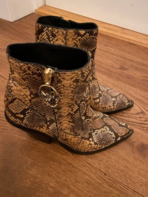 Bruna boots med ormskinnsmönster - Snygga boots med spetsig tå och låg klack i brunt ormskinnsmönster. Skorna har dragkedja på sidan och detaljer i guld. Perfekta för dig som vill sticka ut med ett djuriskt mönster och cool siluett.